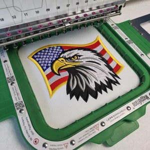 May include: An embroidery machine hoop holding white fabric with a stitched design of a bald eagle head in profile, featuring a yellow beak and black, white, and gray feathers, set against a background of a stylized American flag with red stripes.