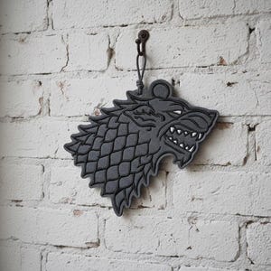 May include: A gray, stylized wolf head wall hanging with a textured surface, hanging from a black cord. The wolf's head is set against a white brick wall.
