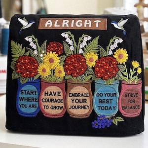 May include: A black fabric cover featuring colourful embroidery of five pots containing flowers. The text reads ALRIGHT, START WHERE YOU ARE, HAVE COURAGE TO GROW, EMBRACE YOUR JOURNEY, DO YOUR BEST TODAY, and STRIVE FOR BALANCE, with two hummingbirds.