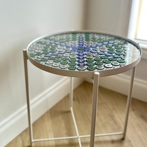 Op de afbeelding: Een ronde bijzettafel met een glazen blad met een mozaïek van groene, witte en blauwe bierdoppen. De tafel heeft een wit metalen frame en poten en staat op een houten vloer. De tafel heeft een diameter van ongeveer 50 cm.
