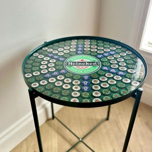 Op de afbeelding: Ronde, groene bijzettafel met een tafelblad van bierdoppen. De tafel heeft een zwart metalen frame en poten. Het Heineken-logo is prominent in het midden van de tafel te zien.