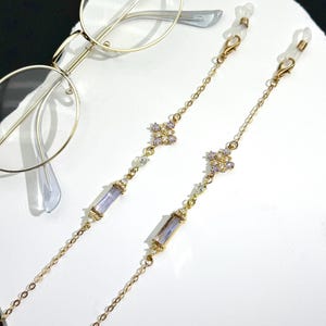 Op de afbeelding: Goudkleurige brillenketting met een delicaat ontwerp. De ketting heeft kleine, heldere en paarse edelstenen en rechthoekige paarse edelstenen. De ketting is ontworpen om een bril vast te houden en wordt getoond naast een bril met gouden montuur.