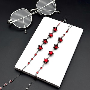 Op de afbeelding: Een bril met een dun zilveren montuur en een decoratieve ketting met donkere metalen schakels, kleine ronde rode kralen en grotere rode stervormige bedels. De ketting ligt over een wit rechthoekig blok en toont het gedetailleerde ontwerp.