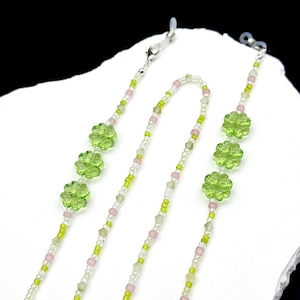 May include: A beaded eyeglass chain featuring translucent light green four-leaf clover charms interspersed with small white, pale pink, and lime green seed beads. The chain includes silver-toned metal clasps at both ends for secure attachment.