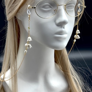 Peut inclure: Une chaîne de lunettes dorée avec des breloques en forme de fleurs de muguet blanches et de fausses perles. La chaîne est attachée à des lunettes à monture dorée sur une tête de mannequin avec des cheveux blonds. La chaîne est conçue pour tenir les lunettes autour du cou.