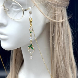 May include: A gold-toned eyeglass chain featuring a delicate pendant with green floral-shaped crystals, clear faceted beads, and a small teardrop-shaped crystal charm. The chain attaches to the glasses with white silicone loops and gold metal clasps.