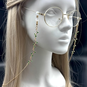 Op de afbeelding: Goudkleurige bril met ronde montuur en een delicate ketting. De ketting heeft een herhalend patroon van kleine groene blaadjes. De bril wordt getoond op een witte mannequinhoofd tegen een donkere achtergrond.