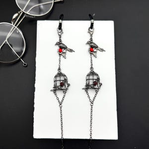 May include: Eyeglass chain with a bird and cage design. The chain features a black bird perched on a red accent, with a cage pendant below. The chain is made of small, linked metal pieces and is designed to hold eyeglasses.