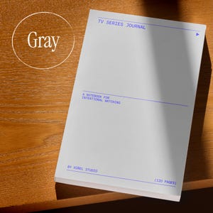 Op de afbeelding: Een grijze notitieboek met de titel "TV Series Journal" met blauwe tekst en lijnen. De kaft leest "A Notebook for Intentional Watching" en "By K&uuml;&ntilde;el Studio". Het notitieboek heeft 120 pagina's en ligt op een houten oppervlak.