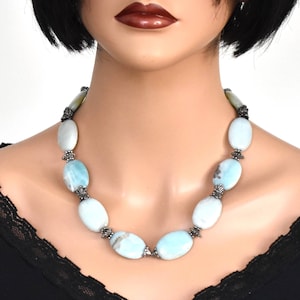 Könnte beinhalten: Eine Halskette mit großen, ovalen, hellblauen und weißen Edelsteinen. Die Steine sind durch verzierte, silberfarbene Metallperlen verbunden. Die Halskette wird auf einer Schaufensterpuppe mit schwarzem Oberteil präsentiert.