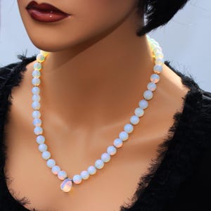 May include: A delicate necklace featuring luminous, round, opalescent beads. The necklace has a teardrop-shaped pendant at the center. The beads have a soft, pearly sheen, and the necklace is set against a black background.