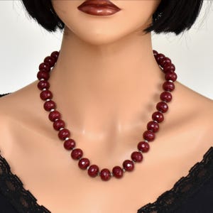 May include: A necklace featuring a single strand of round, faceted, deep red beads. Each bead is separated by a small, metallic silver-toned spacer. The necklace rests against the neck, showcasing a uniform arrangement of the dark red, textured stones.