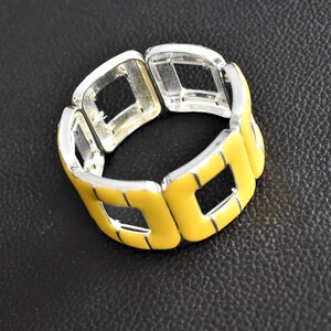 Op de afbeelding: Een elastische armband bestaande uit meerdere zilverkleurige metalen schakels, elk voorzien van een vierkant inlegstuk van felgeel emaille. Het ontwerp bestaat uit herhalende geometrische open segmenten verbonden door elastiek.
