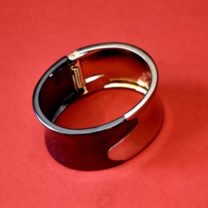 May include: A wide, hinged bracelet with a two-tone design. The bracelet features a dark, glossy finish on the main body and a silver-colored accent. The interior is gold-toned. The bracelet is set against a red background.
