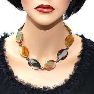 May include: A necklace with large, oval beads in shades of brown, yellow, and green. The beads are connected with silver-coloured metal accents. The necklace is displayed on a mannequin.