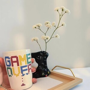 May include: A white ceramic mug with the words "GAME OVER" in pixelated rainbow letters. A black vase holds white flowers, and a wooden tray with a gold handle is in the background.