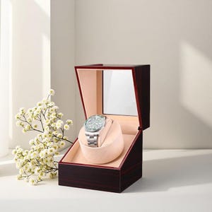 May include: A wooden watch box with a dark brown finish, open to reveal a light pink interior and a silver wristwatch with a green bezel. The box has a mirrored lid and a soft, rounded cushion to hold the watch.