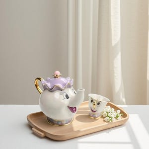 May include: A white ceramic teapot with a purple lid and gold accents, and a matching teacup, both with smiling faces. The teapot and teacup are on a wooden tray, with a small bouquet of white flowers.