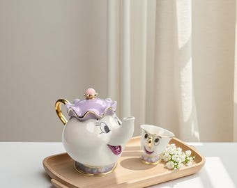 Può includere: Una teiera in ceramica bianca con coperchio viola e dettagli dorati, e una tazza abbinata, entrambe con facce sorridenti. La teiera e la tazza sono su un vassoio di legno, con un piccolo bouquet di fiori bianchi.