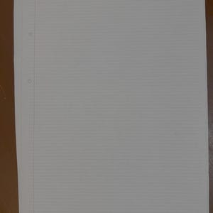 May include: A stack of white, lined A4 paper with three-hole punches along the left edge. The paper has horizontal lines for writing and a vertical margin line. The paper is on a brown surface.