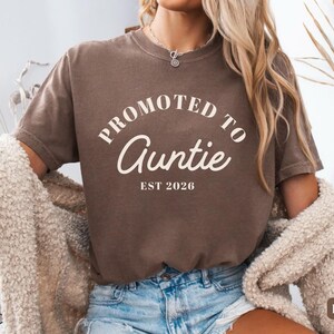 May include: A dark brown short-sleeve t-shirt featuring the cream-coloured text PROMOTED TO Auntie EST 2026 printed on the front. The shirt has a crew neckline and a relaxed fit, paired with light blue denim jeans and a textured beige cardigan.