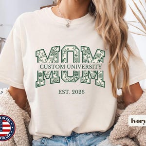 May include: An ivory t-shirt featuring the word MOM in large, dark green block letters filled with a floral pattern. The text CUSTOM UNIVERSITY is printed in the center, with EST. 2026 below the main graphic. The shirt has a crew neckline.