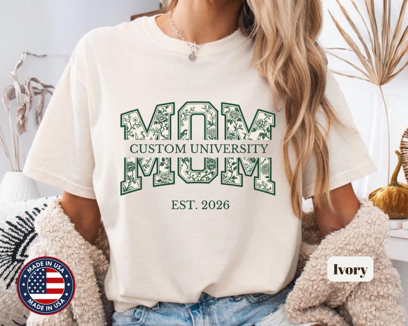 May include: An ivory t-shirt featuring the word MOM in large, dark green block letters filled with a floral pattern. The text CUSTOM UNIVERSITY is printed in the center, with EST. 2026 below the main graphic. The shirt has a crew neckline.