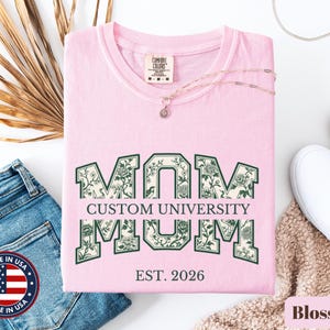 May include: A light pink short-sleeve t-shirt featuring the word MOM in large block letters with a dark green floral pattern. The text CUSTOM UNIVERSITY is printed in the center, and EST. 2026 is printed below. The shirt has a Comfort Colors brand label.