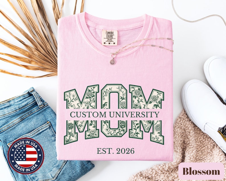 May include: A light pink short-sleeve t-shirt featuring the word MOM in large block letters with a dark green floral pattern. The text CUSTOM UNIVERSITY is printed in the center, and EST. 2026 is printed below. The shirt has a Comfort Colors brand label.