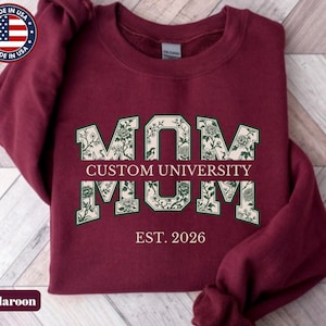 May include: A maroon long-sleeve sweatshirt featuring the word MOM in large block letters with a cream and dark green floral pattern. The text CUSTOM UNIVERSITY is printed in the center, with EST. 2026 below it in a light pink serif font.