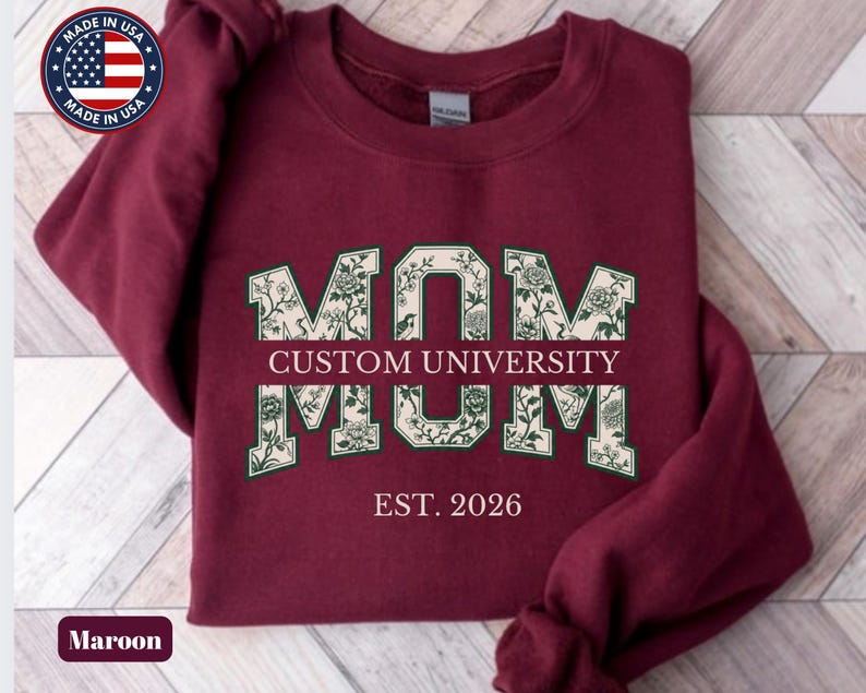 May include: A maroon long-sleeve sweatshirt featuring the word MOM in large block letters with a cream and dark green floral pattern. The text CUSTOM UNIVERSITY is printed in the center, with EST. 2026 below it in a light pink serif font.