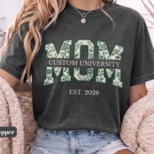 May include: A charcoal gray short-sleeve t-shirt featuring the word MOM in large block letters with a white and green floral pattern. The text CUSTOM UNIVERSITY is printed in a horizontal bar across the center, with EST. 2026 below it.