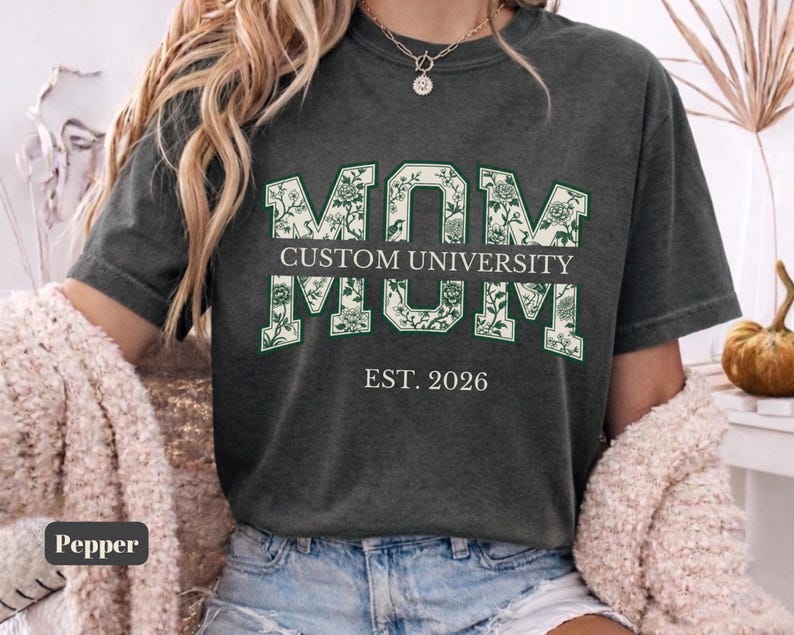May include: A charcoal gray short-sleeve t-shirt featuring the word MOM in large block letters with a white and green floral pattern. The text CUSTOM UNIVERSITY is printed in a horizontal bar across the center, with EST. 2026 below it.