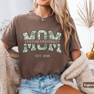 May include: A brown short-sleeve t-shirt featuring the text MOM in large block letters with a green floral pattern. Below the first MOM is the text CUSTOM UNIVERSITY, followed by a second MOM and the text EST. 2026 in a white serif font.
