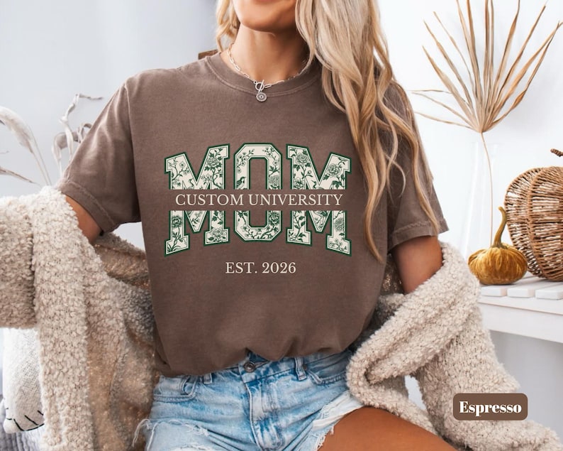 May include: A brown short-sleeve t-shirt featuring the text MOM in large block letters with a green floral pattern. Below the first MOM is the text CUSTOM UNIVERSITY, followed by a second MOM and the text EST. 2026 in a white serif font.