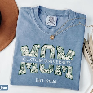 May include: A blue jean colored t-shirt with a crew neckline and a Comfort Colors label. The front features the word MOM in large block letters filled with a green and white floral pattern, with the text CUSTOM UNIVERSITY and EST. 2026.