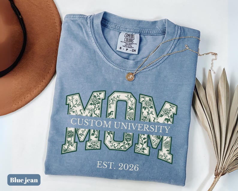 May include: A blue jean colored t-shirt with a crew neckline and a Comfort Colors label. The front features the word MOM in large block letters filled with a green and white floral pattern, with the text CUSTOM UNIVERSITY and EST. 2026.