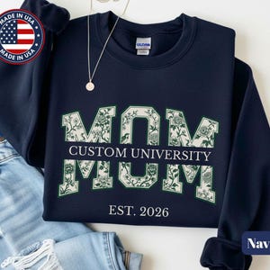 May include: A navy blue crewneck sweatshirt featuring the word MOM in large block letters filled with a white and green floral pattern. A horizontal bar across the center reads CUSTOM UNIVERSITY in white text, with EST. 2026 printed below.