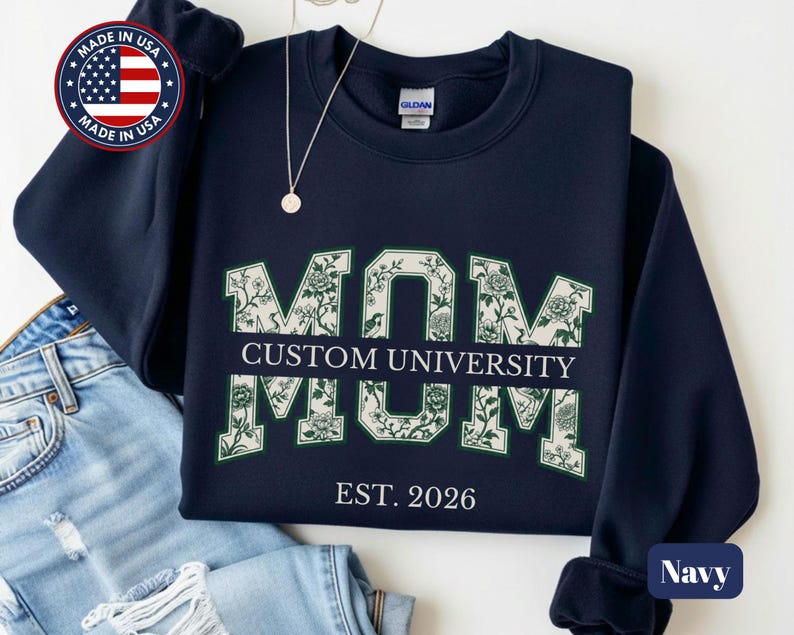 May include: A navy blue crewneck sweatshirt featuring the word MOM in large block letters filled with a white and green floral pattern. A horizontal bar across the center reads CUSTOM UNIVERSITY in white text, with EST. 2026 printed below.