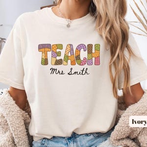 May include: An ivory short-sleeve t-shirt featuring the word TEACH in large, multicoloured, patterned block letters with a patchwork design. Below the word, the name Mrs. Smith is printed in a black cursive script font on the front centre.