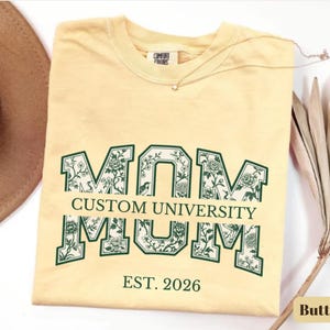 May include: A pale yellow t-shirt featuring the word MOM in large block letters with a dark green outline and a floral pattern inside. The text CUSTOM UNIVERSITY is printed across the center, with EST. 2026 printed in dark green below the design.