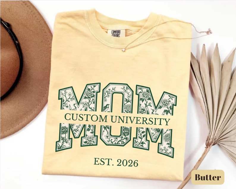 May include: A pale yellow t-shirt featuring the word MOM in large block letters with a dark green outline and a floral pattern inside. The text CUSTOM UNIVERSITY is printed across the center, with EST. 2026 printed in dark green below the design.