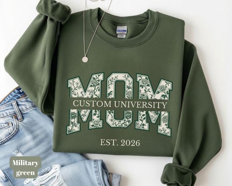 May include: A military green crewneck sweatshirt featuring the word MOM in large block letters with a floral pattern inside. The text CUSTOM UNIVERSITY is centered across the letters, with EST. 2026 printed below. A silver necklace rests on the collar.