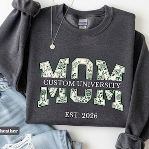 May include: A dark heather gray crewneck sweatshirt featuring the word MOM in large block letters filled with a green and white floral pattern. A horizontal bar across the center reads CUSTOM UNIVERSITY, and the text EST. 2026 appears below.