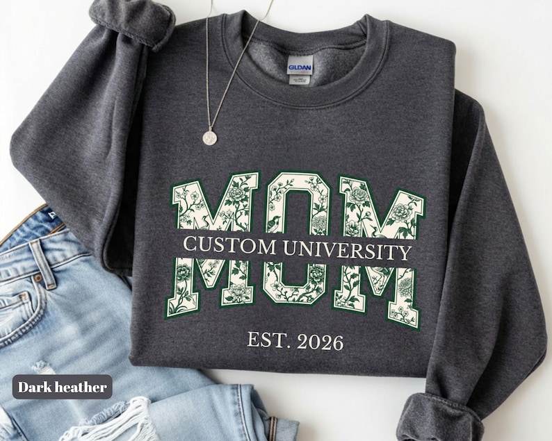 May include: A dark heather gray crewneck sweatshirt featuring the word MOM in large block letters filled with a green and white floral pattern. A horizontal bar across the center reads CUSTOM UNIVERSITY, and the text EST. 2026 appears below.