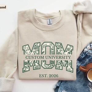 May include: A sand-colored crewneck sweatshirt featuring the word MOM in large block letters with a dark green floral pattern fill. The text CUSTOM UNIVERSITY is centered horizontally across the letters, with EST. 2026 printed in dark green below.
