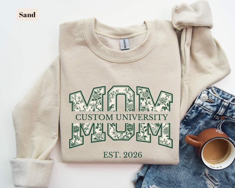 May include: A sand-colored crewneck sweatshirt featuring the word MOM in large block letters with a dark green floral pattern fill. The text CUSTOM UNIVERSITY is centered horizontally across the letters, with EST. 2026 printed in dark green below.