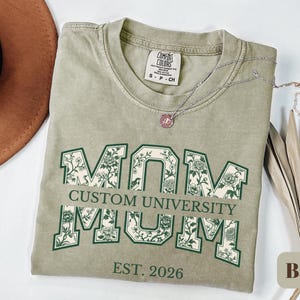 May include: A sage green short-sleeve t-shirt with a crew neckline. The front features the word MOM in large block letters filled with a white and green floral pattern. A horizontal bar across the center reads CUSTOM UNIVERSITY, with EST. 2026 below.