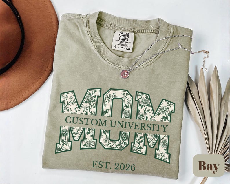 May include: A sage green short-sleeve t-shirt with a crew neckline. The front features the word MOM in large block letters filled with a white and green floral pattern. A horizontal bar across the center reads CUSTOM UNIVERSITY, with EST. 2026 below.