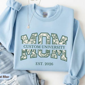May include: A light blue crewneck sweatshirt featuring the word MOM in large block letters with a dark green floral pattern inside. The text CUSTOM UNIVERSITY is printed horizontally across the center, with EST. 2026 printed below in dark green.
